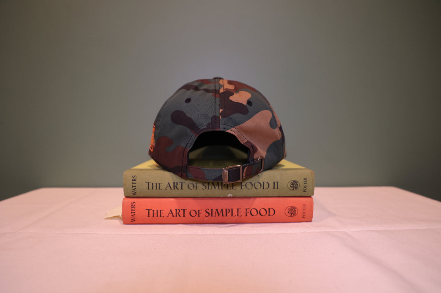 Back view of Camouflage print Baseball hat with an adjustable strap. Hat is on a stack of books titled 'The Art of Simple Food' against a green background.
