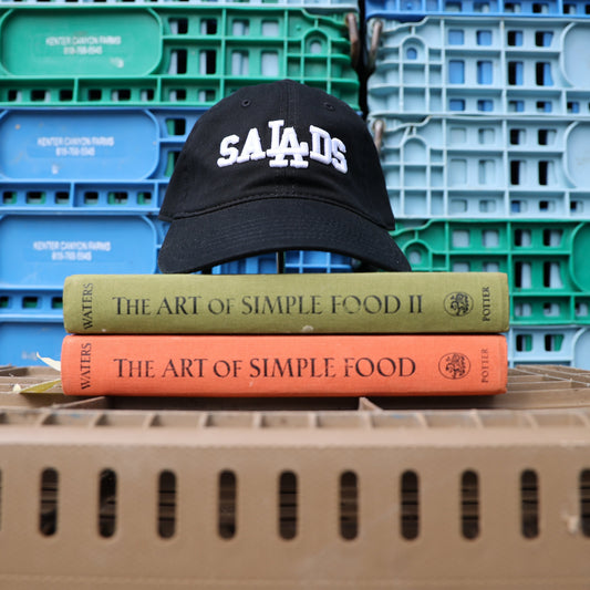Front view of a black baseball cap with 'SALADS' in white embroidered letters sits on top of two hardcover books titled 'The Art of Simple Food' by Alice Waters, placed against a background of stacked blue and green plastic crates.
