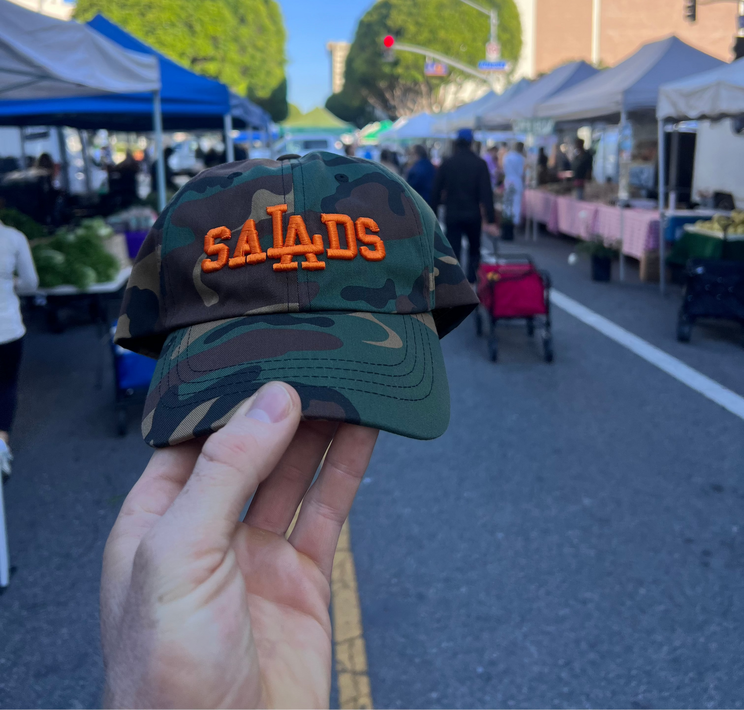 Camouflage cap with 'SALADS' text held up in front of an outdoor market.