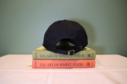 Black cap on top of two books titled 'The Art of Simple Food' with a plain background