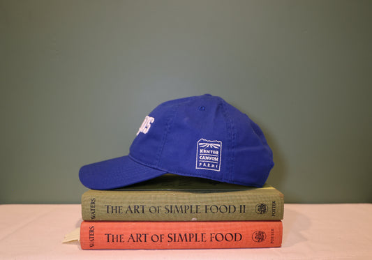Side view of Blue Baseball hat with 'SALADS' text embroidered on the front in white and 'Kenter Canyon Farms' embroidered in white on the left side. Hat is on a stack of books titled 'The Art of Simple Food' against a green background.