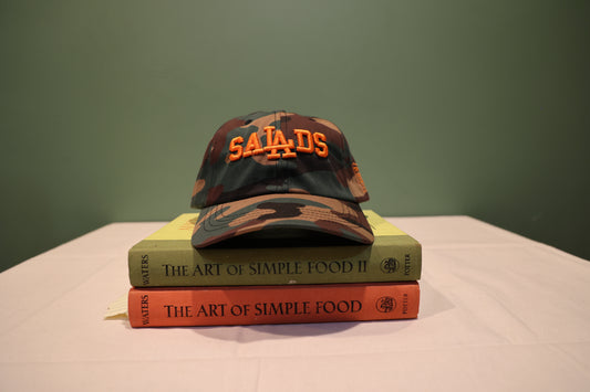 Front view of Camouflage print Baseball hat with 'SALADS' text embroidered on the front in Orange. Hat is on a stack of books titled 'The Art of Simple Food' against a green background.