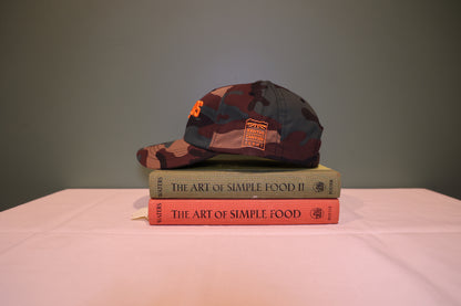 Side view of Camoflauge print Baseball hat with 'SALADS' text embroidered on the front in Orange and 'Kenter Canyon Farms' embroidered in Orange on the left side. Hat is on a stack of books titled 'The Art of Simple Food' against a green background.