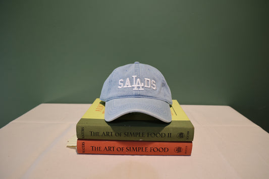 Front view of light-wash denim Baseball hat with 'SALADS' text embroidered in white on the front. Hat is on a stack of books titled 'The Art of Simple Food' against a green background. 