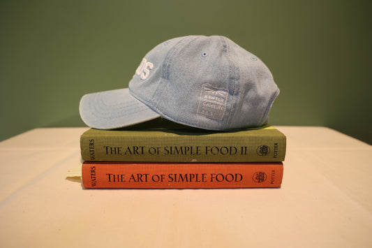 Side view of light wash denim Baseball hat with 'SALADS' text embroidered on the front in white and 'Kenter Canyon Farms' embroidered in white on the left side. Hat is on a stack of books titled 'The Art of Simple Food' against a green background.